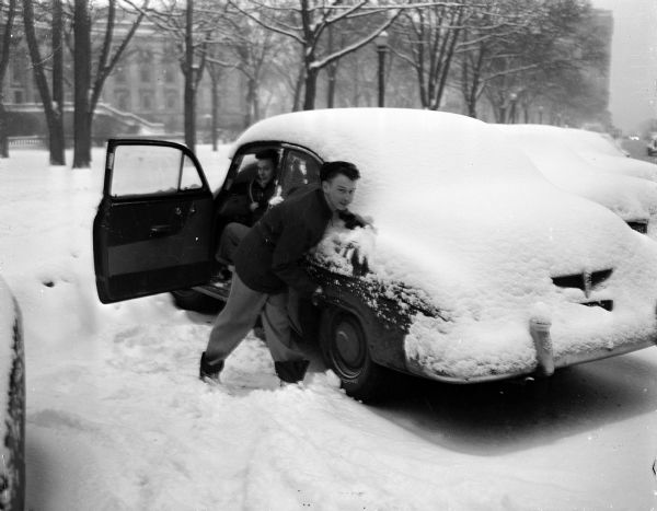 Winter scene with George Schneider and Roy Staley attempting to push a car stuck in the snow away from a curb on the Capitol Square.