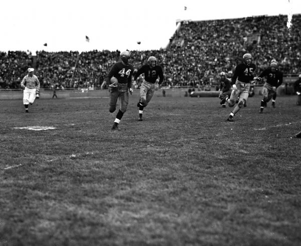 Football players run down the field followed by a referee at the Wisconsin-Iowa football game. The stands are crowded with fans.