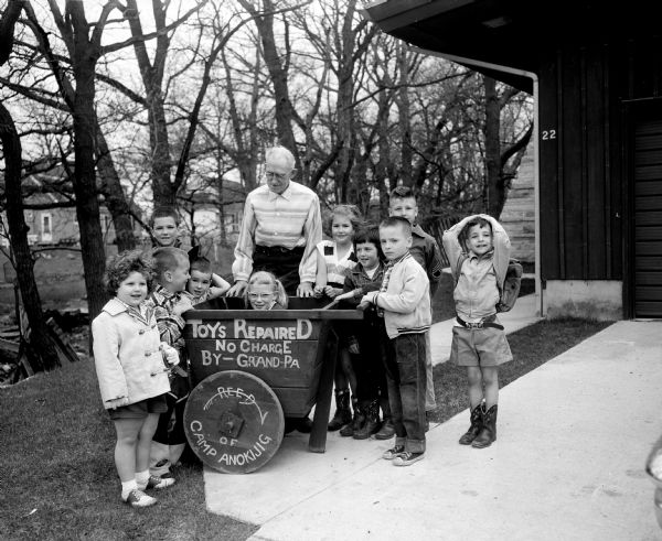 C. Glenn Goodsell is surrounded by children in front of a cart which reads "Toys Repaired No Charge By Grand-Pa". The sign on the wheel of the cart reads: "Reed of Camp Anokijig".