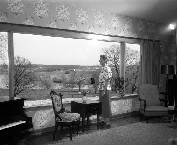 Mrs. Norman Gill is standing and looking out of a window from inside her home at Spring Hill Farm near Cambridge, viewing the surrounding countryside and the Koshkonong river. The Altrusa Club sold (on commission) plants and gardening supplies that were provided by six Madison and Cambridge merchants at the Gill's 80-acre property. Profits from the sale were used for scholarship and hospital funds and plantings for area nursing homes.