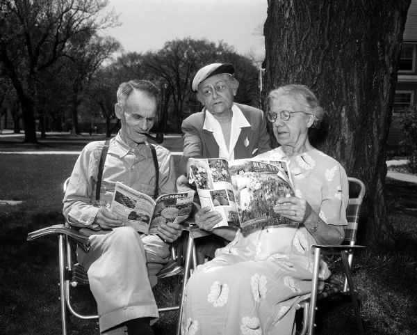 A volunteer discussing seed catalogs with two nursing home residents as they are sitting outside on lawn chairs. They are at Monona Drive Rest Home at 4202 Monona Drive. Sitting are Harry Grounsell and Minnie Gannon. Standing in the middle is volunteer Alfreda Fauerbach. The Altrusa Club raised funds to provide seeds and plantings for Madison area nursing homes.