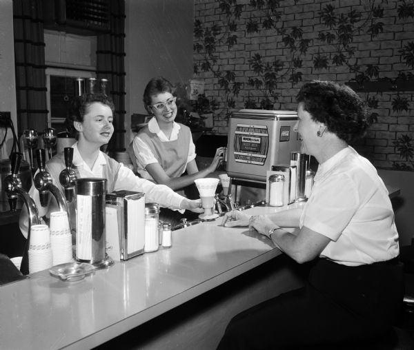 Madison General Hospital Junior Aides Judy Oakland (left) and Karla McCormick (center) serve Frances McPhee (right), member of the hospital auxiliary at the hospital coffee shop/soda fountain.