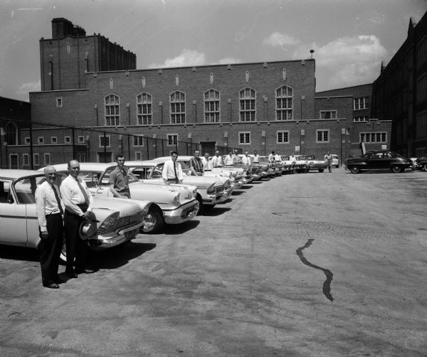 John McDowell (left), supervisor of summer program for Madison public schools, and Milo Swanton, a member of the AAA advisory committee, stand in front of the fleet of dual-control cars to be used in the summer driver education program. A representative of the dealer providing each car stands beside their car. The picture is taken in front of the East High School gymnasium.