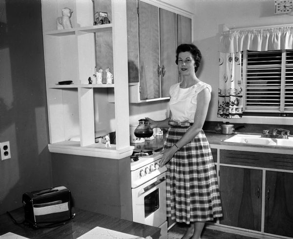 Lois Persen Berres standing in her kitchen while holding a coffee pot. The photograph is one of a series showing how newlywed women have made their new living space attractive and hospitable.