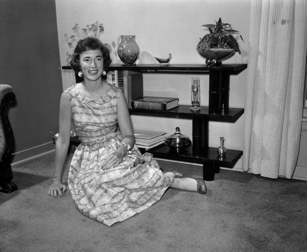 Portrait of Gayle Grelle Cody in the living room of her home, sitting on the floor next to a bookcase that cost two dollars to make. It is made of pine boards and beer cans, varnished and enameled to a high sheen. The photograph is one of a series showing how newly wed women have made their new living space attractive and hospitable.