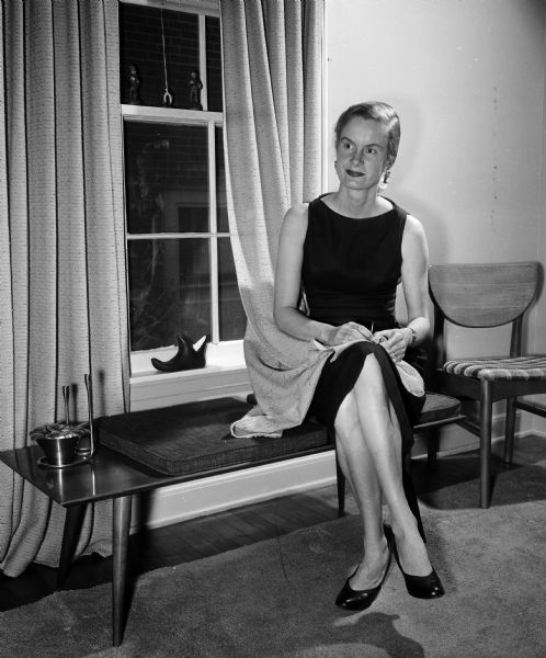 Joanne Peters Rothe sitting in the living room of her home while hemming a drape for a nearby window. The photograph is one of a series showing how newlywed women have made their new living space attractive and hospitable.