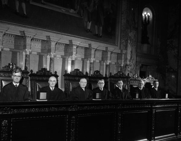 Justices of the Wisconsin Supreme Court are shown posed seated in their court chambers in the Capitol. Justices are, left to right:, E.L. Wingert, George Currie, Grover L. Broadfoot, Chief Justice John F. Martin, Timothy Brown, Roland J. Steinle, and Thomas E. Fairchild.