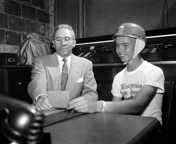 Robert Simpson (right), wearing a soap box derby helmet, receiving a bank book with a $25 savings account as a prize for 'best sport' due to a cut hand and a steering defect disqualification. Herman Kneubuehl (left), First National Bank assistant vice-president, is presenting the bank book. On a shelf behind them are two manual adding machines.