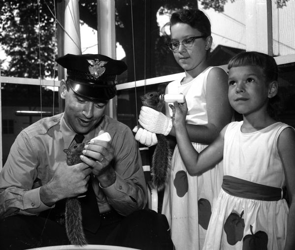 Police officer Edward E. Daley (left) bottle feeding one of three baby squirrels he rescued, while sisters Patty and Cathy Olstadt are looking on and bottle feeding a second squirrel. The negative reveals that the girls are wearing identical (possibly homemade) dresses with decorative sashes and pumpkins.