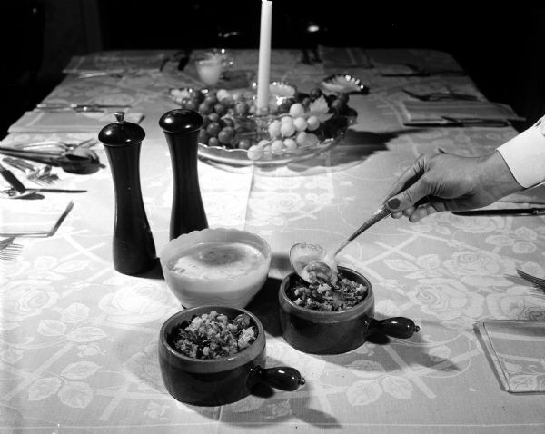 A person's hand holding a ladle is dipping into one of two crocks of seafood casserole (wild rice and fish) on a table decorated with place settings and a center piece.