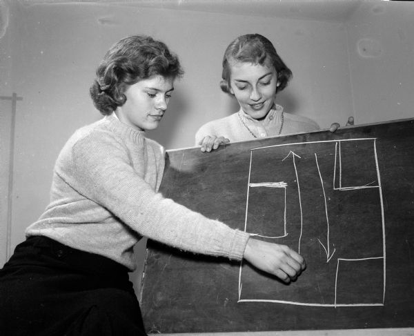 Jean Kleven sketches a diagram of the grand march for the East Side homecoming dance as Delores Klubertanz stands by.
