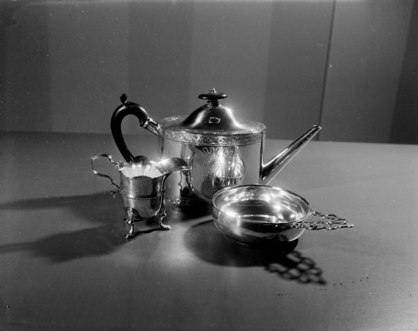 The history of tea service was shown by a display of tea pots in the Wisconsin Historical Society women's auxiliary exhibit, "Tides of Taste." The image shows a 18th century metal tea set consisting of a engraved tea pot, a footed cream dispenser, and a shallow bowl called a porringer often used to hold sugar.