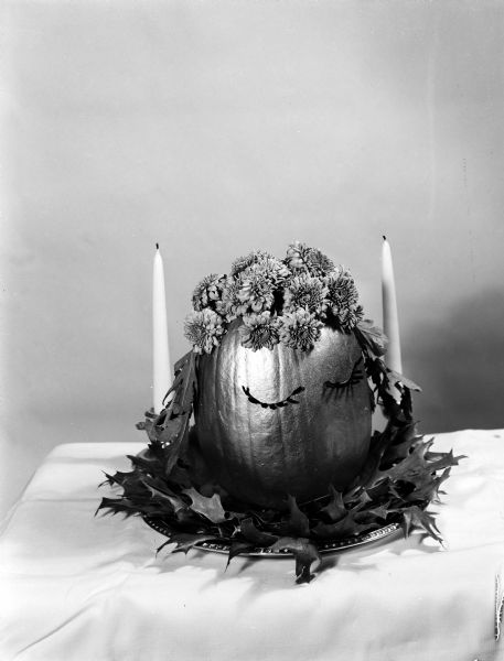 A decorated pumpkin serves as a table centerpiece. It is decorated with a face, a hat made of fresh flowers, and attached leaves for hair.