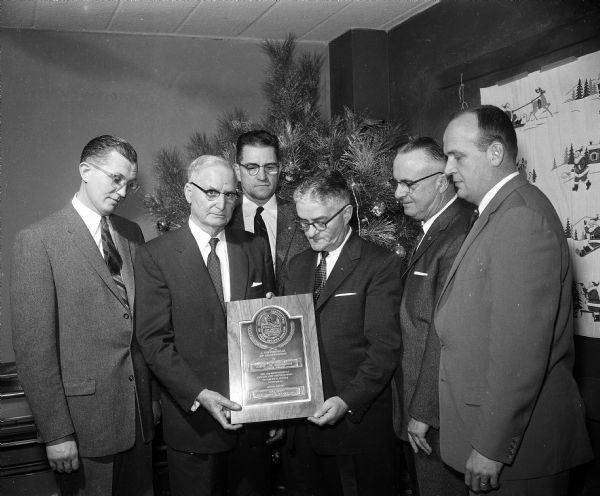 J.S. Miller posing with the plaque he received upon retirement from the Wisconsin Real Estate Brokers board after 14 years service. Shown left to right: M. William Gerrard (La Crosse); J.S. Miller; Julius Dinger (Eau Claire); Jerry Coulter (Madison); Herbert Ganser (West Allis); and Roy Hays (Milwaukee).