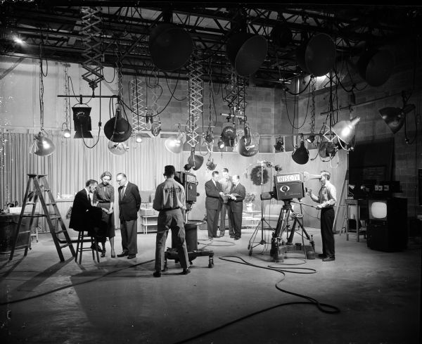 Interior shot of television studio. Large lights are suspended from the ceiling, there are two TV cameras and their operators, a television set, and two groups of three people, all wearing suits. They are preparing for a 15-hour pledge drive for The Wisconsin State Journal's Empty Stocking Club.