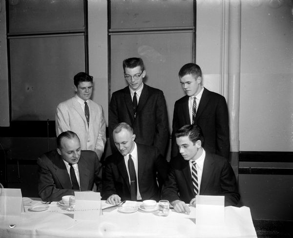 Group portrait of the main speakers at the 36th East High School football banquet. Seated left to right: Herbert (Butch) Mueller, head coach; Rev. Charles Anderson, main speaker; Dick Taylor, tackle who reviewed the season. Standing left to right: Lester Nesbit, captain and toastmaster; Hugh (Pat) Richter, who spoke on 1958 prospects; and Jerry Nickles, who reviewed the Four Lakes League.