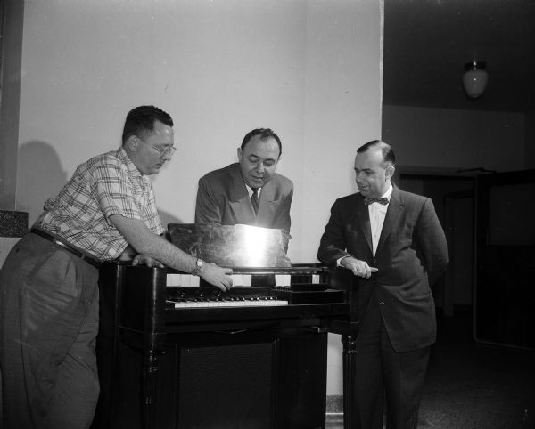 Three men examine an electric organ placed upon a wheeled platform. The caption states, a new portable chord organ for patients at Mendota State Hospital absorbs the interest of Wilton Duckworth. State executive director for mental health, Dr. Walter Urben, superintendent of the hospital, and John McNamara, hospital music director.  The organ is a gift of the Mental Health Associations of Dane, Grant, and Eau Claire counties. It will increase the amount of entertainment provided for patients.