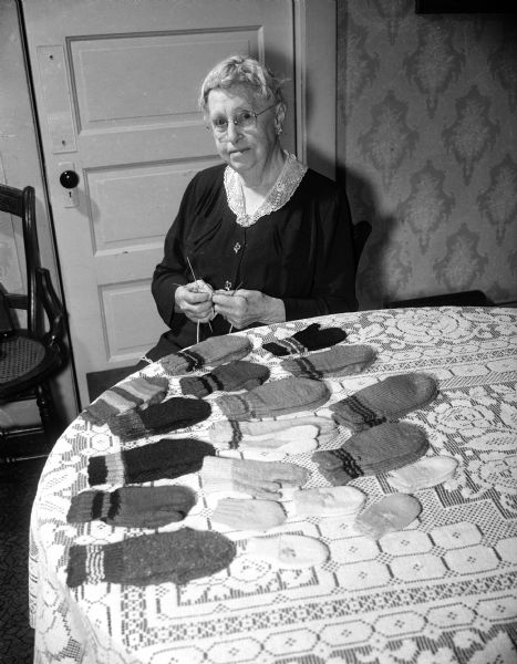 Mary Gillin is sitting behind a table with a display of 21 pairs of mittens of various sizes with wrist stripes that she knitted to donate to The Wisconsin State Journal's Empty Stocking Club.