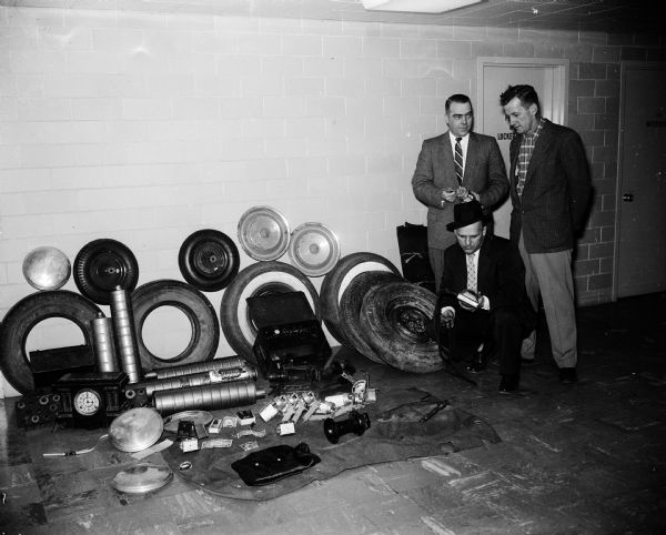 The original caption states: "Dane County sheriff's officials survey about $300 worth of auto parts and other merchandise recovered from members of a juvenile theft gang at Cambridge. Sheriff Fred Goff, left, Deputy John Zeller and Deputy Earl Sorenson, foreground, said they believe the gang includes about 20 members who have stolen at least $1,000 worth of items in six months."