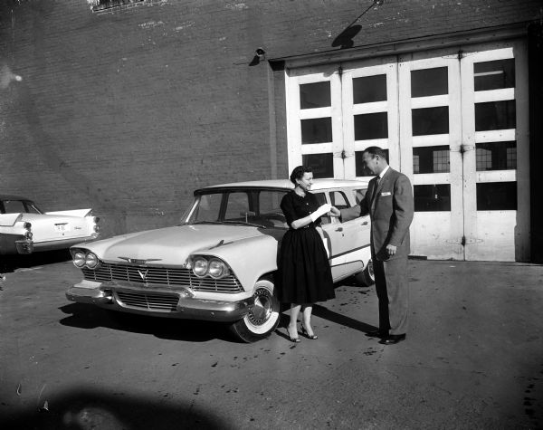A man hands keys to a woman (possibly Mrs. Howard Radder) while standing in front of a Plymouth station wagon at Gillespie-Blumer Motors, 802 East Washington Avenue. Mrs. Howard Radder was possibly a contest winner.