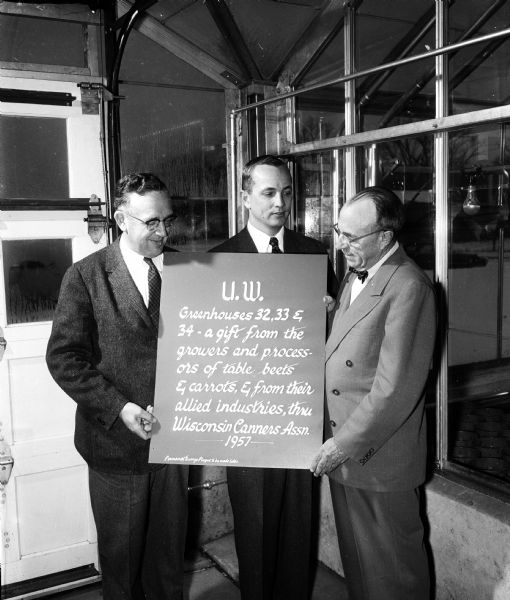 Three men are shown holding a large copy of the wording to be put on a bronze plaque to mark the ceremony of turning a 100-foot greenhouse over to the University of Wisconsin. The greenhouse was given by the Beet and Carrot Growers and Processors of the Wisconsin Canners Association.