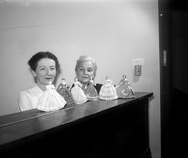 Leonora Courtenay and Marjory Bush stand behind a shelf upon which six dolls dressed in hand-made authentic costumes of 1858 are posed. The dolls were a part of the fashion show and tea sponsored by the Women's Auxiliary of Grace Episcopal Church to celebrate the congregation's centennial.