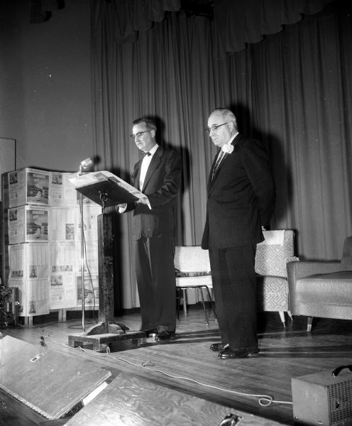 Jack Burke standing at a podium onstage while reading a tribute and life story of Armand F. Ketterer, principal of Franklin school, standing at right.