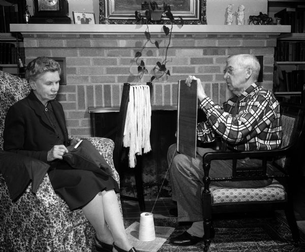 Mr. W.J. Haight and his wife Charlotte of Cambridge are sitting in front of their fireplace. Mr. Haight, chairman of the Cambridge Community Red Cross Drive, cuts lengths of yarn to be used for a Red Cross project. Charlotte, organizer of a Red Cross production and sewing unit, knits a "ditty bag" for servicemen.