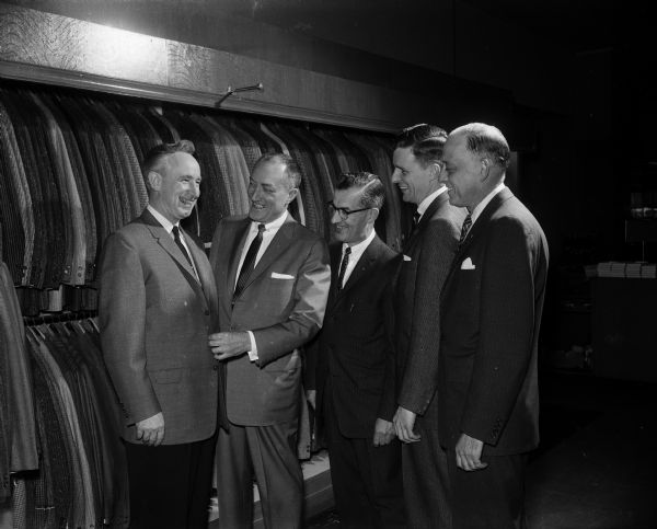 Governor Vernon W. Thomson (left) trying on a new suit, as four men representing four men's clothing stores, all located on the Capitol Square, are looking on. The salesmen's names and clothing stores are, left to right, E.W. Holmquist (Spoo and Son), Adolf Knocke (Karstens), Robert Schmitz (The Hub), and Jack Aycock (Olson and Veerhusen).  'Dress Right Week' was sponsored by the National Institute of Men's and Boy's Wear.