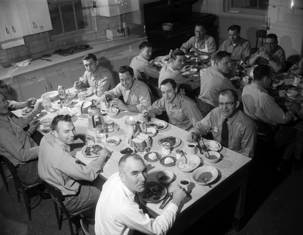 Elevated view of 13 firemen sitting at two tables, ready to eat their 11 am meal. The men are at Central Station at 18 South Webster Street.  From left to right at the far table, facing camera, are Phil Behrend (the cook), Percy Moen, and Gilman Stone. Opposite them are, left to right, Charles Merkle, Don Berray, Robert Mutch, and Peter Vallem. At the front table, second row, are William Block, Dick Healy, Joe Lawrence, and Harlie Lippolt. In front are Donald Wilder and (station chief) William Lynaugh.