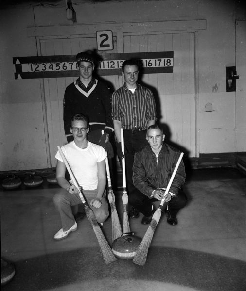 Portrait of four members of the Wisconsin High School Dave Strobel Rink team. The men are holding four brooms near the curling stone in the foreground. Kneeling are Perry Schultz and Stewart Quisling. Standing are Strobel (wearing a tam) and Ted Winans. They earned second place in the Madison high school curling tournament.