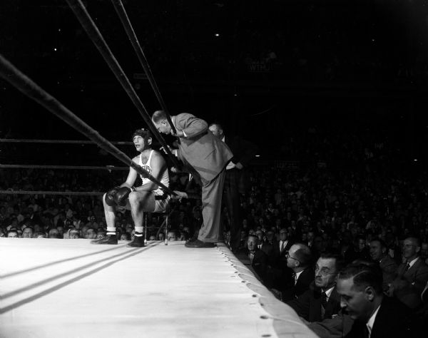 University of Wisconsin heavyweight boxer Ron Freeman is resting in his corner while John J. Walsh is leaning over the ropes with close-up coaching instruction. It is the final bout of the final tournament of Walsh's 25 year coaching career. The right side reveals a large audience, with many men wearing suits.