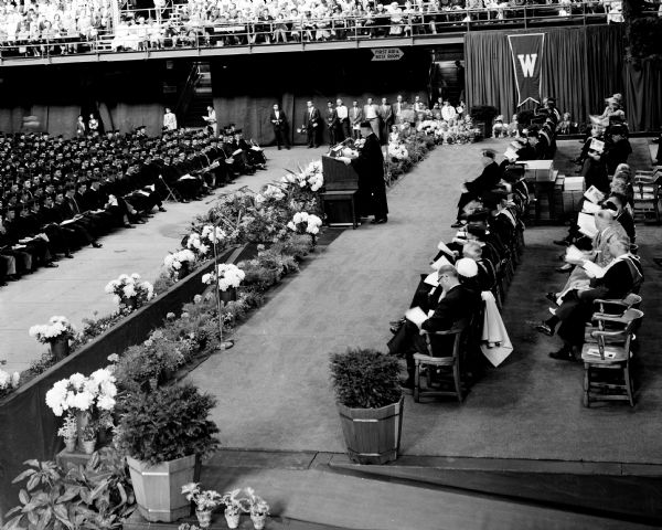 View of the University of Wisconsin's graduation ceremonies as seen from the balcony overlooking the side of the stage. Special guests are seated on the stage while a speaker stands at a podium. Many graduates in caps and gowns are seated on the auditorium floor.
