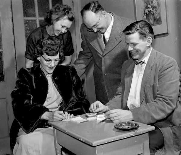 The original caption states: "Foster parents from Dane County met for a get-together sponsored by the foster home committee of the Community Welfare Council.  Registering with Lee Smook, seated right, of the Lutheran Welfare society, are three foster parents, Mrs. Albert Beloungy, seated; Mrs. Herbert Milestone; and Mr. Beloungy. The Beloungys, who have five children of their own, have one foster child, and Mrs. Milestone has two deaf foster children."
