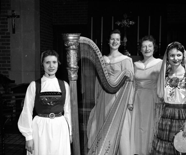 Four women in ethnic costume and gowns pose behind a harp. Shown left to right are: Marjunice West, who sang songs of Norway and is shown in a dress which belonged to her great-grandmother, the late Mrs. R.N. Qualley; harpists Vicki Vauk and Mrs. Cooper; and Elizabeth A. Risser (Mrs. Fred A. Risser), who played rhythm numbers and wore a costume brought from a Caribbbean cruise.