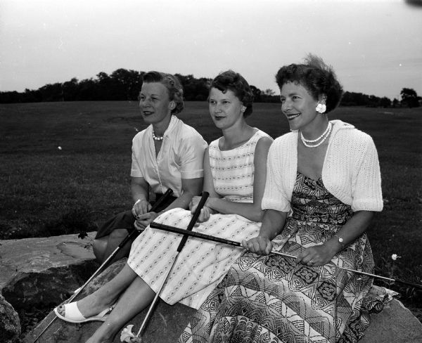 Doris Bailey, Virginia Semrad, and Elizabeth Bruns holding golf clubs while posing on a bench at a golf course. The women are wearing dresses and necklaces.