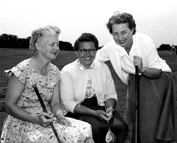 Three women are holding golf clubs and wearing dresses while posing on a bench on a golf course fairway. The original photograph caption states: "The Women's Municipal Golf Association held its annual luncheon at the new clubhouse at the West Side park municipal golf course. The women, of course, played golf before the luncheon." The women are Ruth Holcomb, co-chairman for the luncheon; Mary Keller; and Ruth Nedderman, social chairman.