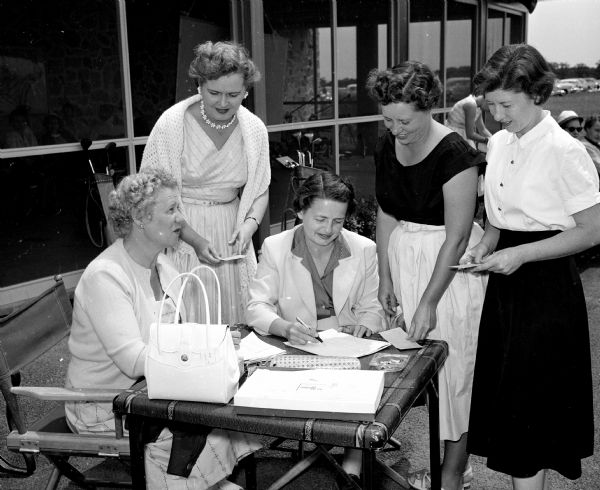 Five women golfers gather around a folding table to add up their scores for the day. The club house, with a large screened porch, is behind them. They are, left to right: Viola O'Neil, hostess; Helen Schmidt, co-chairman of handicaps; Virginia Wrabetz, hostess; Anna Zeman, co-chairman of pairings; and Janie Gilbert, pairings chairman.