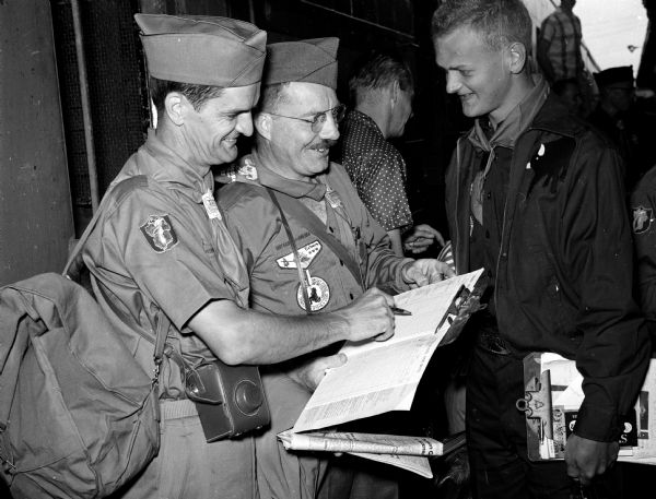 Three men at the railroad station examine a schedule as they leave for the Boy Scout Jamboree in Valley Forge, Pennsylvania. Wallace Wikoff of the <i>Wisconsin State Journal</i> editorial staff is shown with Jamboree-bound participants Art Wolrath (Madison) and Peter Antles (Portage).