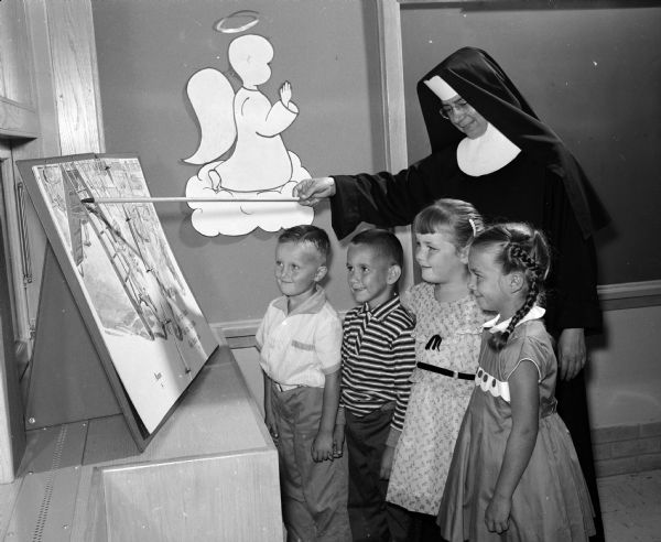 Four first graders look on as Sister Mary Cabrini uses a pointer to draw their attention to a large picture. The children are Michael Crary, William Leute, Dennise Marquis, and Ellen McQueeney. The school had 1,655 pupils from the Westport, Waunakee, and Fox Bluff area.