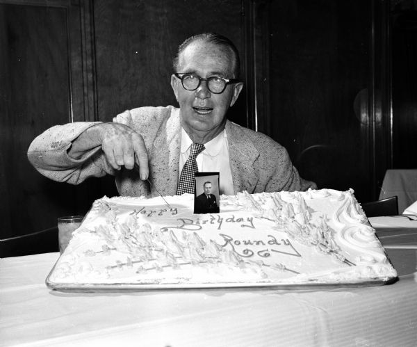 Joseph (Roundy) Couglin, a popular sports columnist for the <i>Wisconsin State Journal</i>, cutting a large decorated birthday cake at his birthday party.