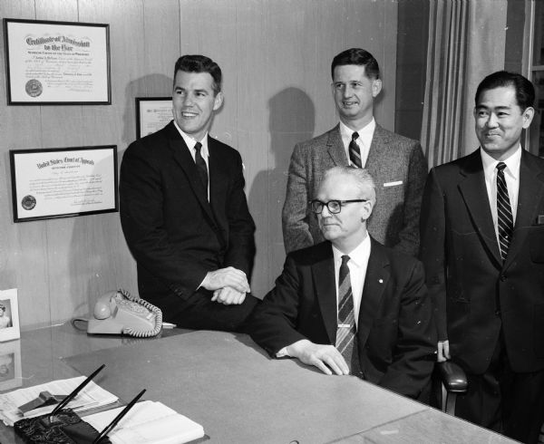 Four men pose for a portrait in an office. Second from the left, standing, is Bruce Thomas. A law degree for Arthur A. McLeod hangs on the wall.