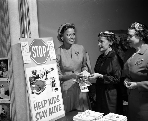 Three women attend an event to display Dane County Medical Society Women's Auxiliary projects. They include, from left to right, Ina Marlow, Emily Sprague, and Lucille Fosmark. They look at a poster focused on the group's interest in safety education.