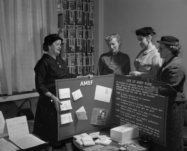 Four women attend an event to display Dane County Medical Society Women's Auxiliary projects. Agnes Hill (left), Helen M. Wasserberger, Beatrice Kabler, and Jeanne L. Parkin look at a display that depicts how contributions of $12,000 to the AMEF (Academy of Medicine Education Foundation) fund have been spent.