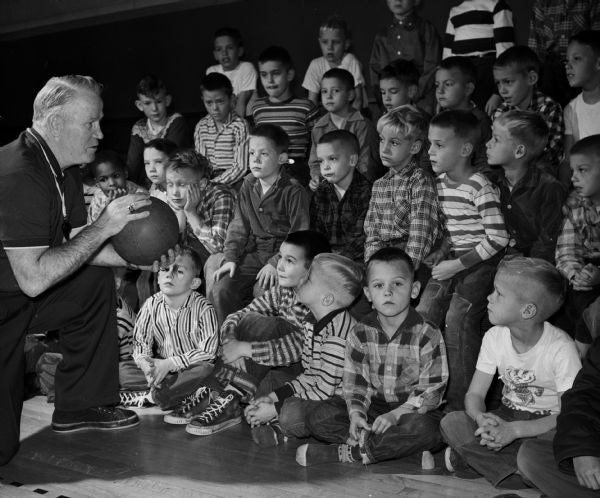 Vern Woodward (left), U.W. Madison's assistant boxing coach and director of the Little Badgers program, talking to one of his classes of more than 25 little boys. The program was in its 20th year and was increasingly popular; it consisted of instruction in boxing, basketball, body conditioning, and group games. They are meeting at St. Bernard's Grade school gymnasium.