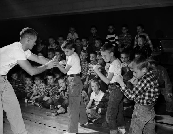 Glenn Nording (left), Middleton High School basketball coach and former U.W. Wisconsin boxer, corrects the boxing postures of three boys who are holding up their fists. Behind them, a group of more than 30 little boys are sitting and watching. The program was in its 20th year and was increasingly popular; it consisted of instruction in boxing, basketball, body conditioning, and group games.
