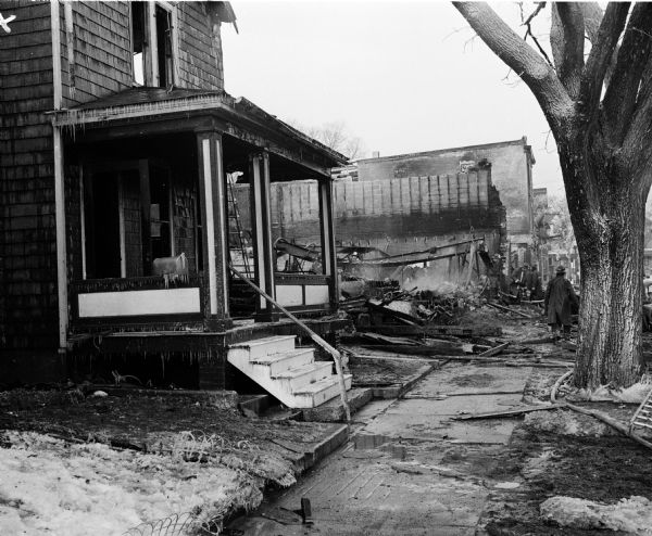 View of a badly damaged house at left, and a pile of rubble in the background where the home of William and Gladys McGrath once stood. Both McGraths were killed in the blast and fire. This was one of two separate explosions and fires that took place on North Bassett Street and East Main Street. The Bassett Street event also destroyed Hyland Hall & Co., White Cross Pharmacy, and Capitol Upholstering Shop. The Main Street event destroyed Hansen Auto Body Works and portions of the Red Arrow Sales Co., and a quonset hut nearby.