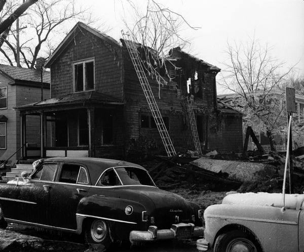View of the badly damaged house at left, and a pile of rubble at right where the home of William and Gladys McGrath once stood. Both McGraths were killed in the blast and fire. This was one of two separate explosions and fires that took place on North Bassett Street and East Main Street. The Bassett Street event also destroyed Hyland Hall & Co., White Cross Pharmacy, and Capitol Upholstering Shop. The Main Street event destroyed Hansen Auto Body Works and portions of the Red Arrow Sales Co., and a quonset hut nearby.