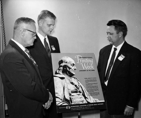 Lawrence E. Perry (left), and Robert Perry, both of Watertown, and Mark Widen, Madison, look at a poster about Printing Week during a dinner observing International Printing Week.