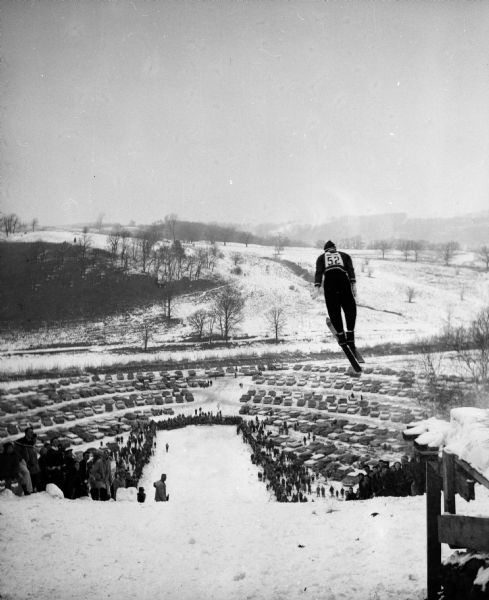 Ski jumper Dale Severson of Eau Claire is shown (center right) flying and leaning high over the Tomahawk Ridge ski jump. Spectators and parked cars are in the background.